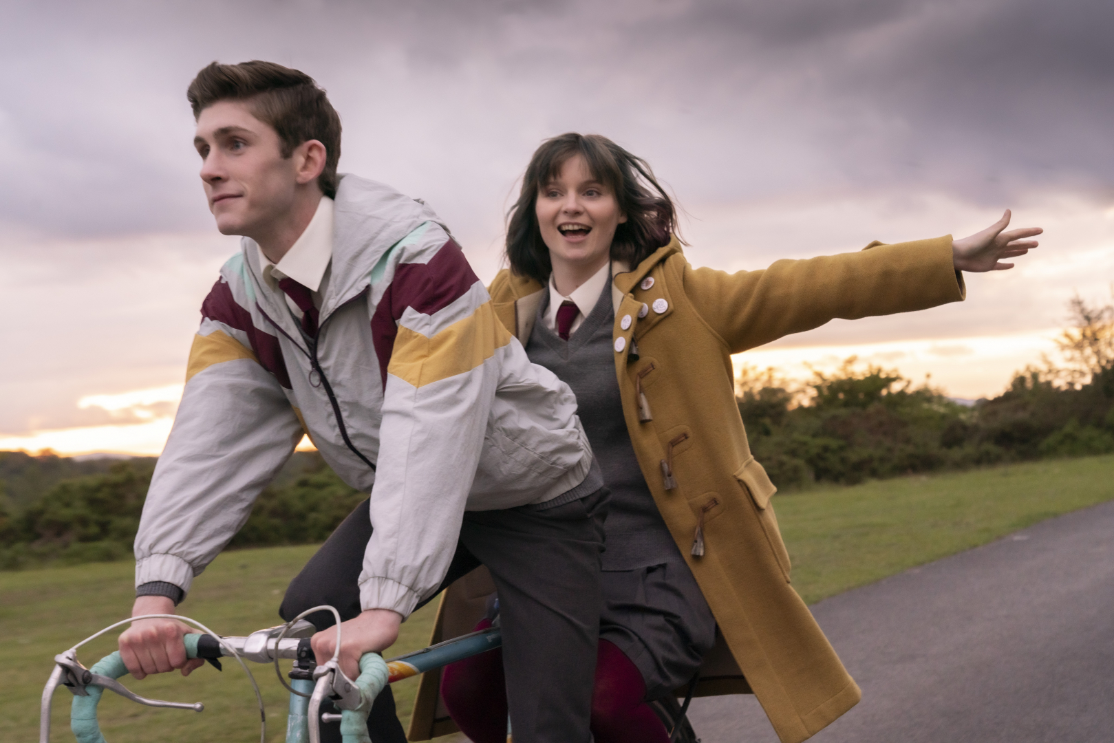 A still from Dating Amber. A worried looking teen boy is riding a bike with a teen girl spreading her arms happily behind him.