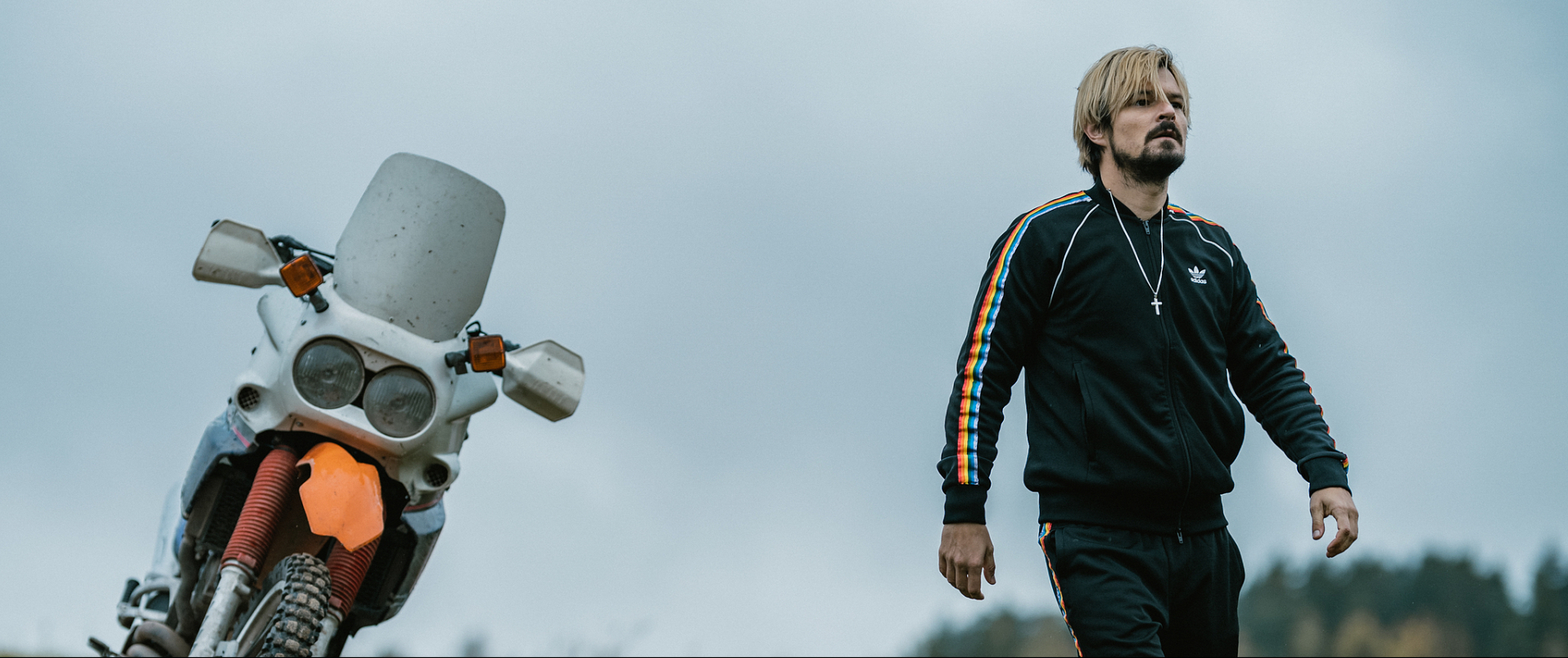 A still from All Our Fears. A man is walking away from a motobike. He is wearing an adidas tracksuit with pride flag stripes on the side and a large cross necklace.
