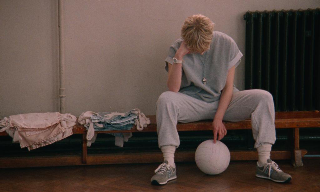 A still from Blue Jean. Jean is sitting on a bench in a school gym and looking down at a ball. She is white woman with short blond hair wearing gray eighties sports clothes.