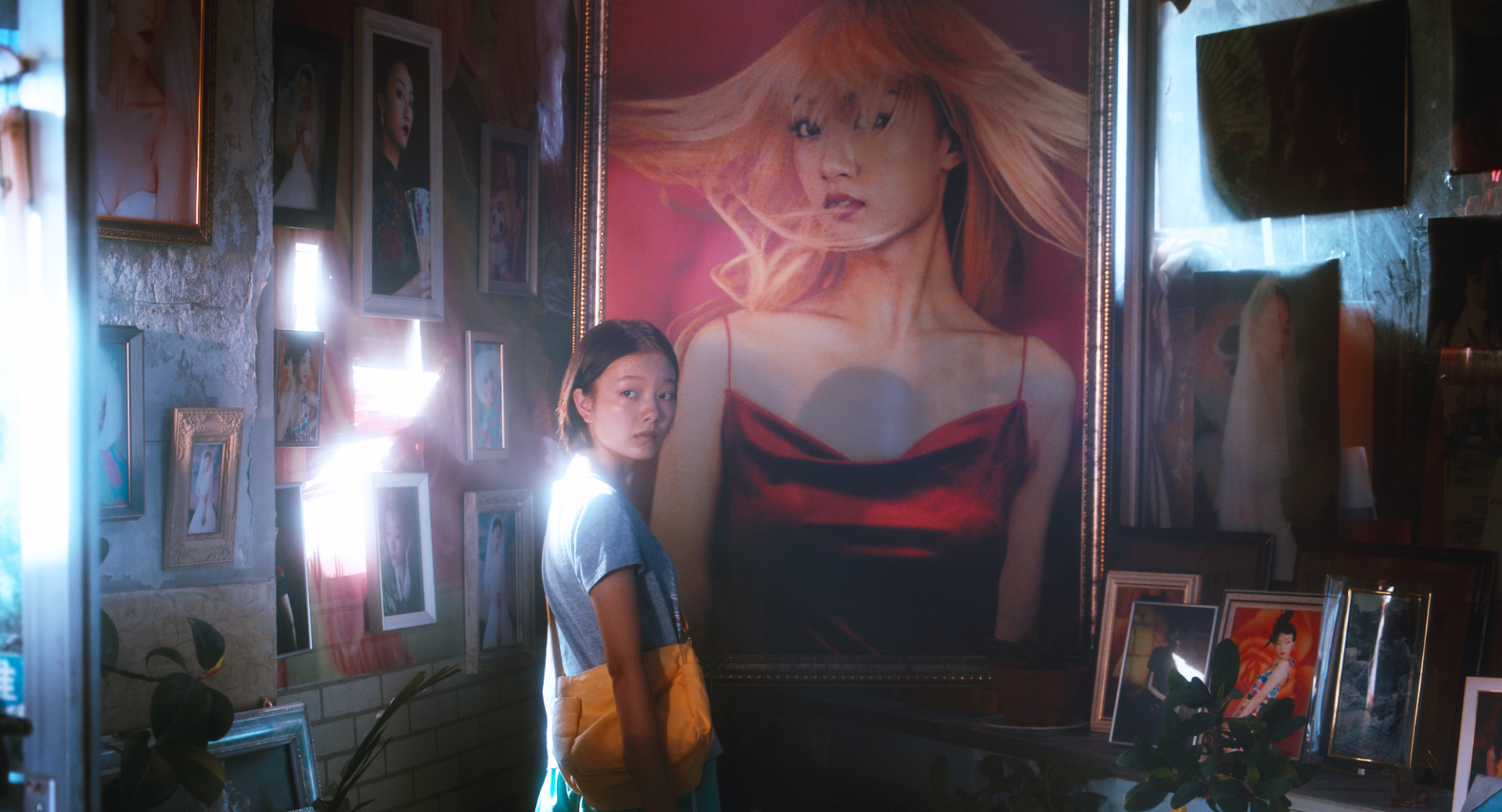 A still from A Song Sung Blue. Liu Xian, a young Chinese girl, is standing in front of many different photographs, including a large one of a woman wearing a blonde wig. The lighting is very luminous.